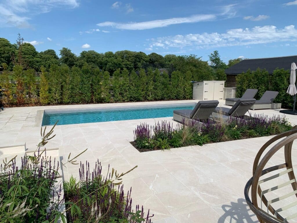 Outdoor swimming pool area with light stone paving, sun loungers, and purple flower beds